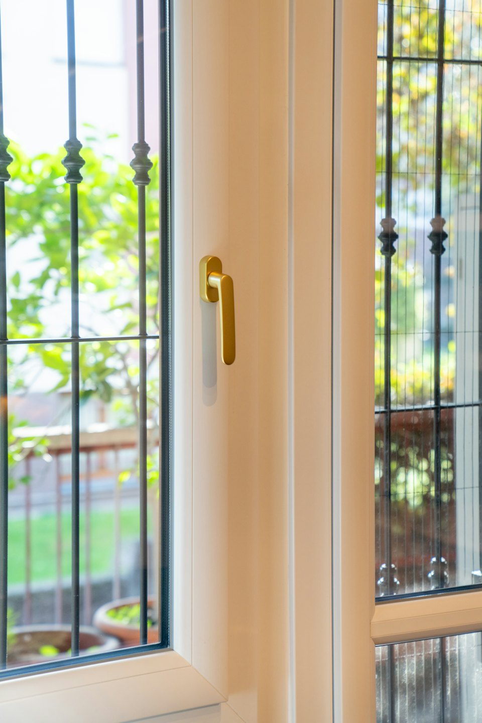 Moderner weißer Fensterrahmen mit goldenem Griff und Blick auf grünes Blattwerk draußen.